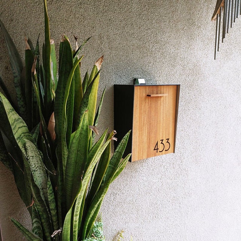May include: A modern black and wood mailbox with the number 433 on the front. The mailbox is mounted on a light gray wall with a green plant in the foreground.