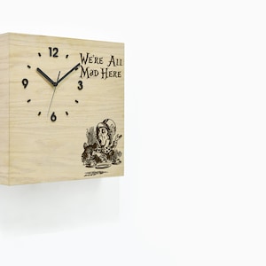 Puede incluir: Un reloj cuadrado de madera con manecillas negras y una ilustración en blanco y negro del Sombrerero Loco de Alicia en el País de las Maravillas. La esfera del reloj dice "We're All Mad Here".