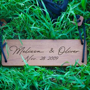 May include: A wooden swing with the names "Melissa & Oliver" and the date "Nov. 28 2009" engraved on it. The swing is hanging from black ropes and is resting on green grass.