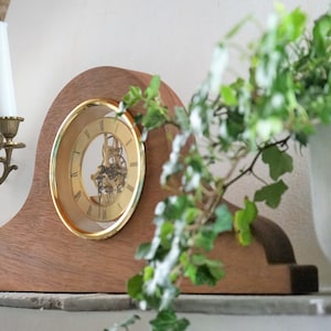 Op de afbeelding: Een houten mantelklok met een gouden wijzerplaat en een zichtbaar uurwerk. De klok staat op een plank met een messing kandelaar en een plant in een pot.