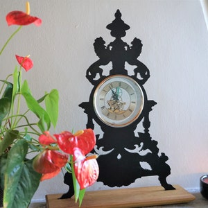 May include: A black metal clock with an ornate design, featuring a round clock face with Roman numerals. The clock is on a wooden base and is surrounded by a red flower.