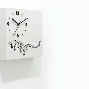 May include: White square clock with black hands and numbers. The clock face has a design of musical notes flowing across the bottom. The clock is mounted on a white wall. The text "Time Box" is in the lower right corner of the image.