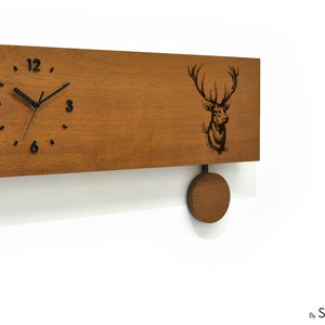 May include: A wooden wall clock with a pendulum. The clock face has black Roman numerals and hands. The clock is decorated with a laser-engraved image of a deer head. The clock is made by Sol Pixie Dust.