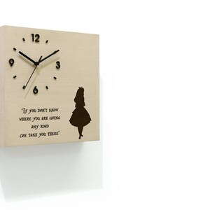 Puede incluir: Un reloj de pared cuadrado de madera con manecillas y números negros. El reloj presenta la cita "Si no sabes a dónde vas, cualquier camino te llevará allí" y una silueta de Alicia en el país de las maravillas.