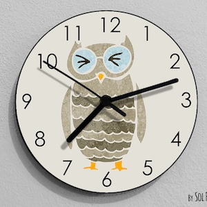 May include: A white round clock with black numbers and hands. The clock features a cartoon owl with brown and grey feathers, blue eyes, and a yellow beak. The text "BY SOL PIXIE DUST" is printed at the bottom of the clock.