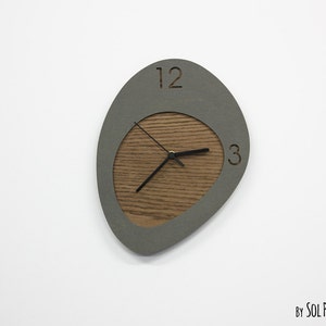 May include: A gray and wood wall clock with a unique oval shape. The clock face has a wood grain texture and the numbers 12 and 3 are visible.