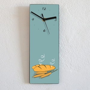 Puede incluir: Un reloj de pared azul claro con agujas negras y números romanos negros. La esfera del reloj tiene un diseño de tres barras de pan con espigas de trigo. El reloj está colgado en una pared blanca.