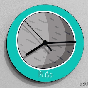 以下が含まれることがあります： ターコイズ色の丸い時計で、グレーと白の冥王星の画像が描かれています。時計の針は黒で、画像の下には「Pluto」という文字が黒で印刷されています。時計の下部には「By SOL PIXIE DUST」という文字が黒で印刷されています。