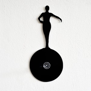 May include: Black silhouette of a person standing on a black circular disc. The person is in a standing pose with arms outstretched.