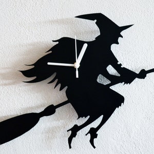 Op de afbeelding: Zwarte silhouet wandklok in de vorm van een heks die op een bezem vliegt. De klok heeft witte wijzers en een zwarte achtergrond.