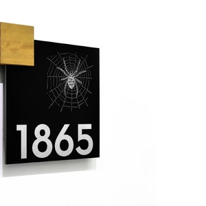 May include: Black square house number sign with a white spider web and spider design and the number 1865 in white. The sign is mounted on a white wall with a small wooden block attached to the top corner.