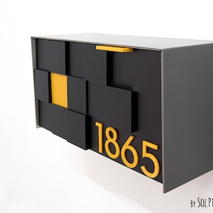 Może przedstawiać: Nowoczesna czarna skrzynka pocztowa o geometrycznym wzorze, z żółtymi akcentami i numerem 1865. Skrzynka ma prostokątny kształt i jest zamontowana na białej ścianie. Wymiary: około 40 cm x 25 cm x 15 cm.