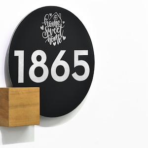 May include: Black circular sign with the number 1865 in silver lettering and the text "home sweet home" in white lettering with a small house icon and hearts. The sign is mounted on a wooden block.