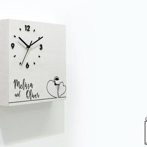 May include: White wooden cuckoo clock with black hands and numbers. The clock face has a black outline of two hearts. The clock is personalized with the names "Melissa and Oliver".