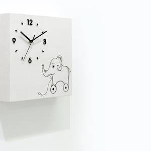May include: White square clock with black hands and numbers. The clock face features a black and white illustration of a toy elephant with wheels.