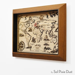 May include: Wooden world map art in a shadow box frame. The map is etched with continents, oceans, and illustrations of ships and compasses. The frame is a warm brown color, and the map has red pins marking locations.