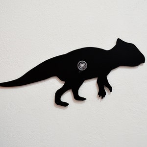 May include: Black silhouette of a dinosaur, mounted on a white wall. The dinosaur is facing right and has a long tail. The silhouette is made of metal and has a small hole in the center for hanging.