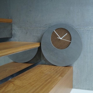 May include: A round, gray concrete clock with white hands and a wood accent on the face. The clock is mounted on a wall.