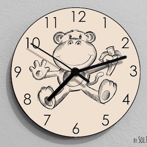 May include: A cream-colored clock with black numbers and hands features a cartoon monkey holding a banana. The monkey is drawn in black and white and has a happy expression. The clock face reads "By Sol Pixie Dust".