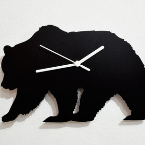 Puede incluir: Un reloj de pared negro con forma de oso y manecillas blancas. El reloj es una silueta de un oso en posición de caminar. El reloj está montado en una pared blanca.