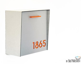 May include: A rectangular silver metal mailbox featuring a bright orange handle at the top and the orange house number 1865 displayed prominently on the front panel. The mailbox has a clean, flat design with a smooth, metallic finish.