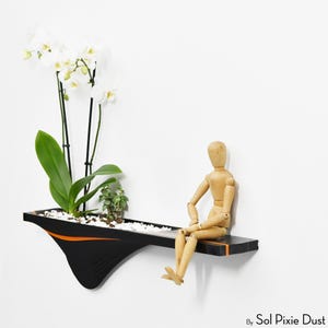 Könnte beinhalten: Ein schwarzer und orangefarbener Wandtopf mit weißen Kieselsteinen, der eine Orchidee und kleine grüne Pflanzen enthält. Eine Holz-Künstlermodell sitzt am Rand. Der Pflanztopf hat ein einzigartiges, modernes Design, ideal für die Präsentation von Pflanzen in Innenräumen.