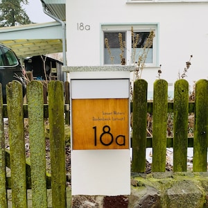 May include: A white mailbox with a wooden panel displaying the text "Sunset Waters Rodenbeck (privat)" and the number "18a". The mailbox is attached to a white post and is in front of a green wooden fence.