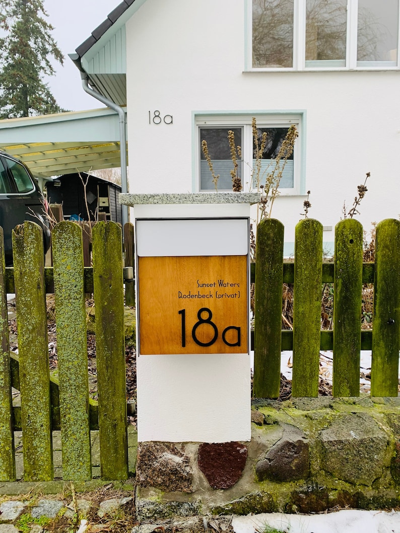 May include: A white mailbox with a wooden panel displaying the text "Sunset Waters Rodenbeck (privat)" and the number "18a". The mailbox is attached to a white post and is in front of a green wooden fence.