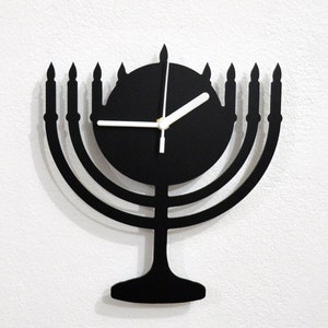 May include: Black menorah-shaped wall clock with a white clock face and hands. The clock is made of metal and has a minimalist design.
