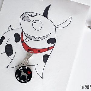 Könnte beinhalten: Ein schwarz-weißer Cartoon-Hund mit einem roten Halsband und einem schwarzen Anhänger, auf dem "Maza" und eine Telefonnummer stehen. Der Anhänger ist an einem silbernen Schlüsselanhänger befestigt.