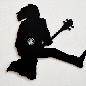 May include: Black silhouette of a person playing a bass guitar, the silhouette is cut out of metal and mounted on a white wall.