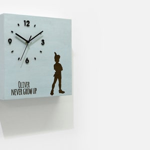Puede incluir: Un reloj cuadrado de madera azul claro con agujas negras y números negros. El reloj tiene una silueta de Peter Pan con el texto "Oliver Never Grow Up".