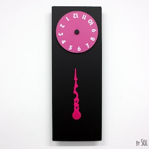 Peut inclure: Une horloge rose et noire avec un pendule unique de forme abstraite rose. Le cadran de l'horloge a des chiffres blancs sur un fond rose.