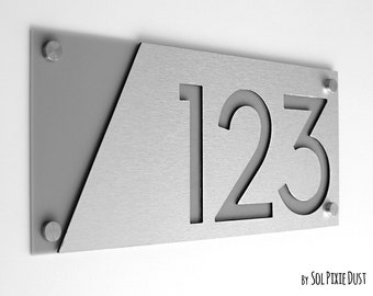 Peut inclure: Une plaque de numéro de maison moderne avec le numéro 123 découpé dans un panneau en métal argenté brossé. L'arrière-plan est un panneau en acrylique gris. La plaque est fixée avec quatre vis argentées.