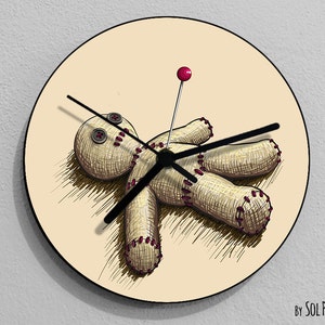 May include: A beige round clock with a black clock face and hands. The clock face features a hand-drawn illustration of a voodoo doll with a red pin in its chest. The doll is lying on its back with its arms outstretched. The clock face is surrounded by a beige border.