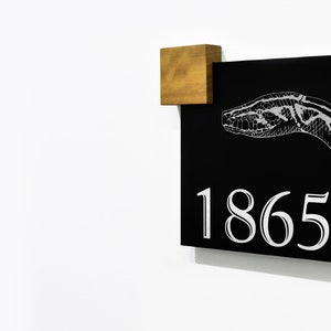 May include: A black sign with a white snake illustration and the number 1865. A small, square wooden block is attached to the top left corner of the sign. The sign is mounted on a white wall.