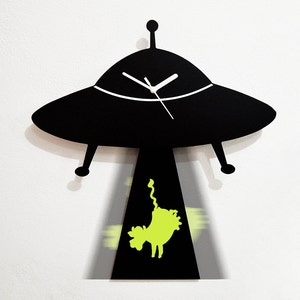 May include: Black UFO-shaped clock with a white clock face and black hands. The clock is hanging on a white wall and is casting a shadow of a green alien figure.