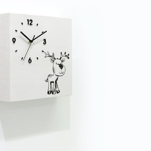 May include: White square clock with black hands and numbers. A black and white illustration of a reindeer is on the clock face. The clock is mounted on a white wall.