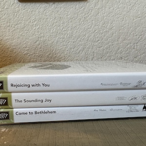 May include: Three white Stampin' Up! stamp set cases stacked horizontally. The top case is labeled Rejoicing with You, the middle case is labeled The Sounding Joy, and the bottom case is labeled Come to Bethlehem, each featuring black text and graphics.