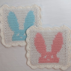 May include: Two crocheted bunny-shaped squares with scalloped edges. One square is white with a light blue bunny, and the other is white with a pink bunny.