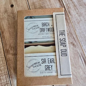 Peut inclure: Une boîte rectangulaire en carton brun contenant deux pains de savon étiquetés The Soap Duo. Le savon du haut est Birch + Driftwood avec bouleau blanc, ambre et brume marine, poids net 130g. Le savon du bas est Sir Earl Grey avec thé noir, bergamote et musc, poids net 130g.