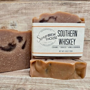 Peut inclure: Trois savons artisanaux de couleur marron et beige. Un savon est empilé sur deux autres. L'étiquette indique "SOUTHERN WHISKEY" avec les ingrédients "CARAMEL * TOBACCO * VANILLA BOURBON". Le poids net est de 130g.