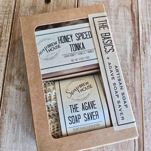 Peut inclure: Une boîte cadeau en carton brun contenant un savon Honey Spiced Tonka et un porte-savon en agave texturé. L'étiquette du savon indique Soap Brew House, Honey Spiced Tonka, Dark Honey, Vanilla, Amber, 130g. L'étiquette du porte-savon indique The Agave Soap Saver.