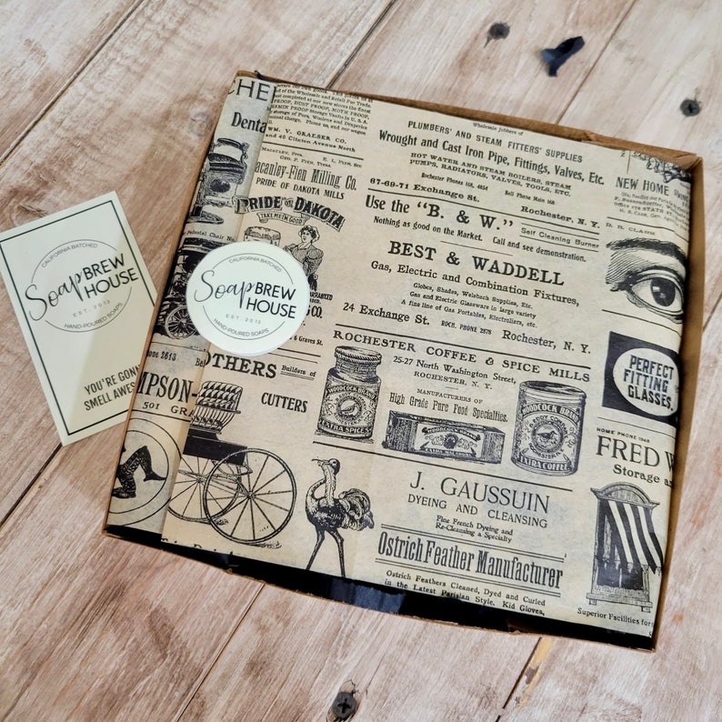 Peut inclure: Une bo&icirc;te en carton marron avec un design de journal vintage. La bo&icirc;te est remplie d'un savon rond blanc avec le texte "Soap Brew House" imprim&eacute; dessus. Le design du journal pr&eacute;sente des illustrations en noir et blanc d'un oiseau, d'une cal&egrave;che et d'une bo&icirc;te de caf&eacute;.