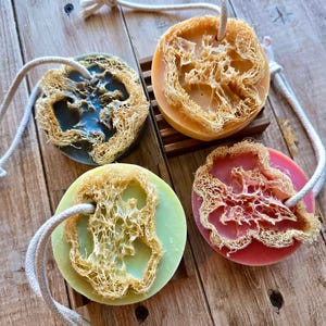 May include: Four round soaps in shades of green, pink, yellow, and blue, each with a natural loofah sponge attached. The soaps have a rope loop for hanging. The soaps are on a wooden surface.