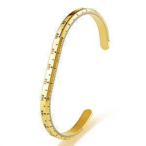 May include: A gold-toned metal bracelet with a ruler design. The bracelet is open and has a curved shape.