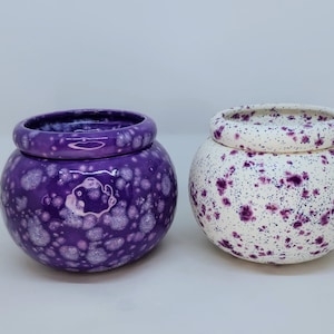 May include: Two ceramic, round pots. One is a deep purple with a speckled pattern, the other is white with purple speckles. Both have a matching lid and a glossy finish. The pots are likely for decorative use.