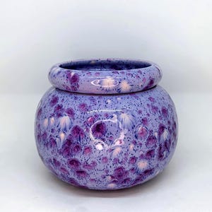 May include: A round ceramic planter with a wide, thick rim and a bulbous body. The surface features a glossy lavender glaze with a crystalline pattern of dark purple and white starburst-like spots scattered across the entire exterior.