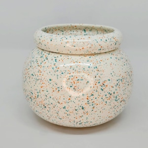 May include: A speckled ceramic jar with a lid. The jar is off-white with a pattern of teal and orange speckles. The lid is a ring shape that sits on top of the jar. The jar has a rounded shape.