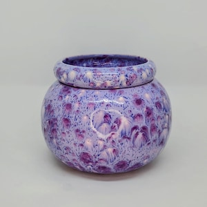 May include: A round ceramic planter with a wide, thick rim and a bulbous body. The surface features a glossy glaze in shades of light lavender and purple, decorated with a speckled, mottled pattern of darker purple and white crystalline blooms.